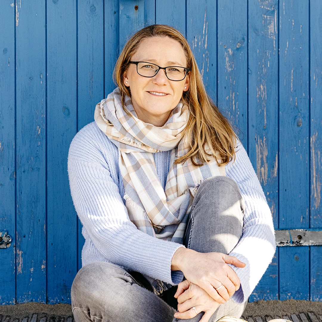 Sarah Evans sitting infront of a blue beach hut holding one knee